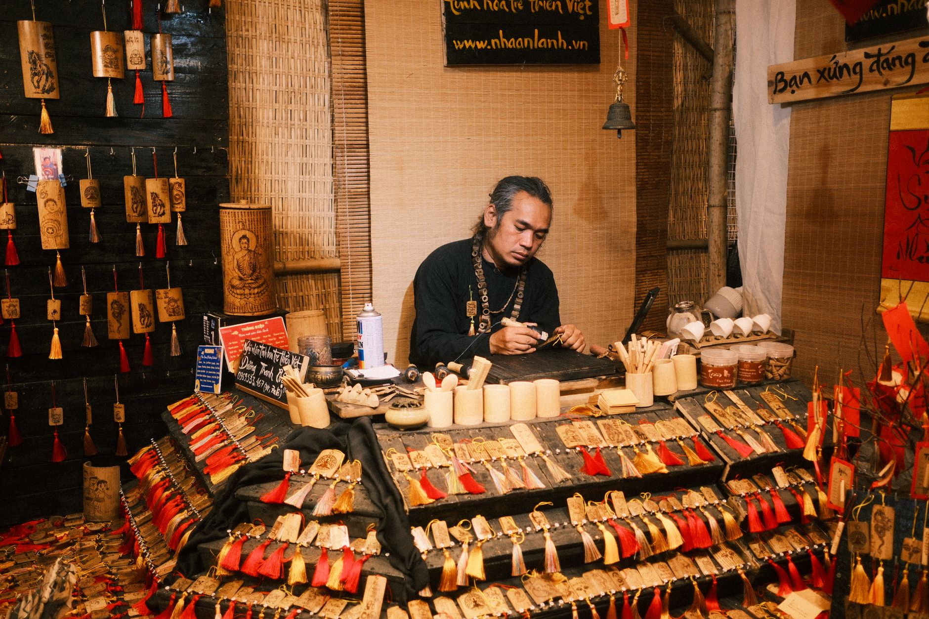Artisan assis derrière son stand de marché, en train de travailler une pièce en bois, présentoir garni de petits objets en bois et étiquettes